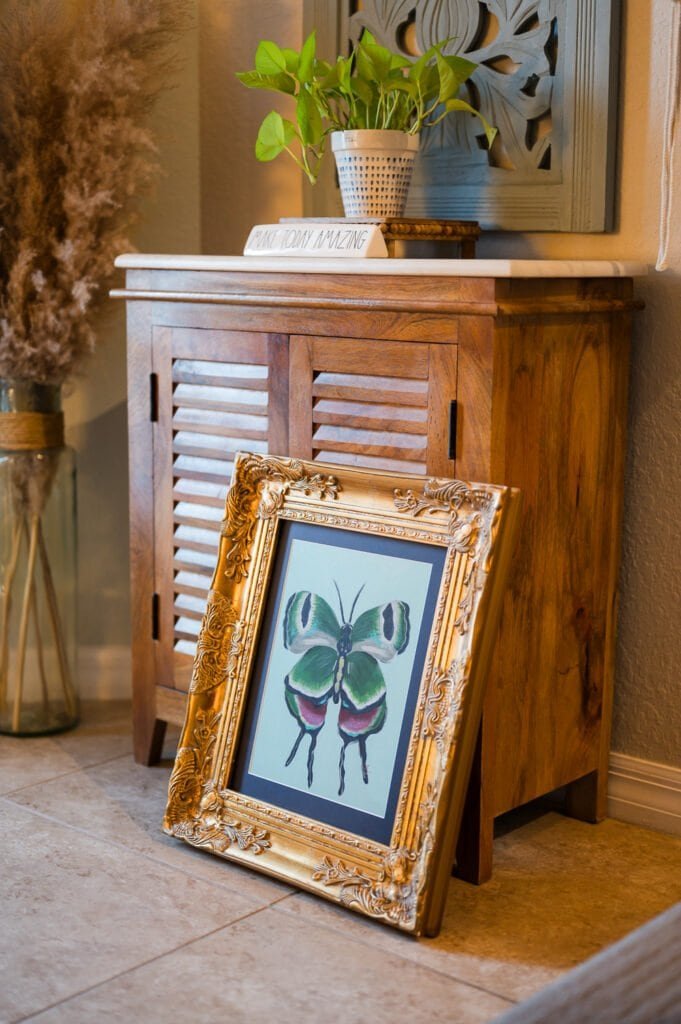 Framed butterfly artwork leaning against a wooden cabinet in a cozy interior space.