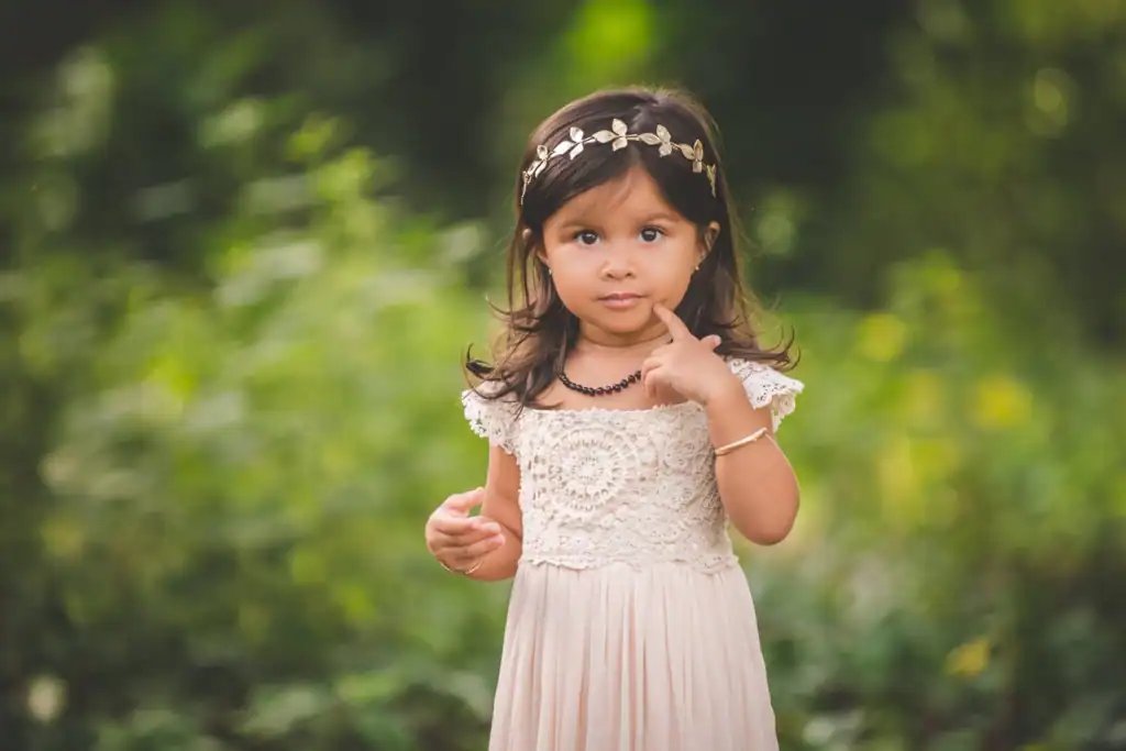 Young girl in a cream dress with a floral headband standing outdoors in a lush green setting.