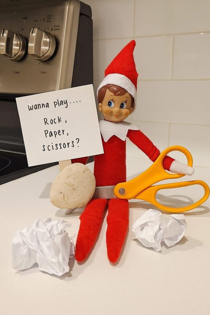 Elf on the Shelf holding a rock, scissors, and paper balls for a rock-paper-scissors setup. Simple two-minute elf idea.