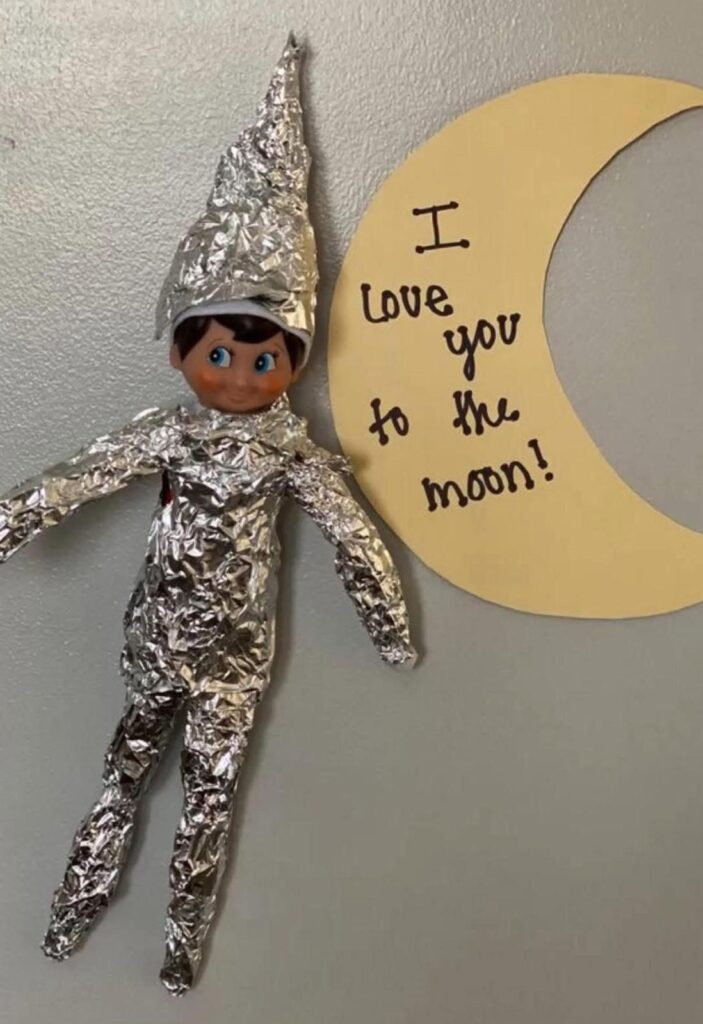 Elf on the Shelf dressed as a foil astronaut next to a paper moon with the message “I love you to the moon.” Easy space-themed elf idea.