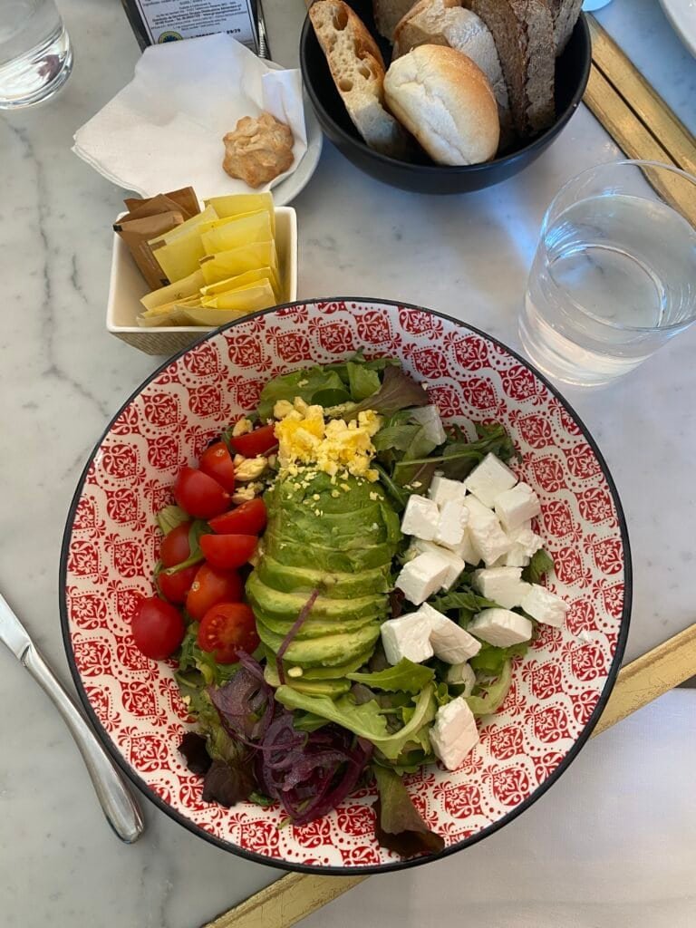 A vibrant, colorful salad featuring cherry tomatoes, sliced avocado, feta cheese cubes, and mixed greens, served in a decorative red and white patterned bowl.