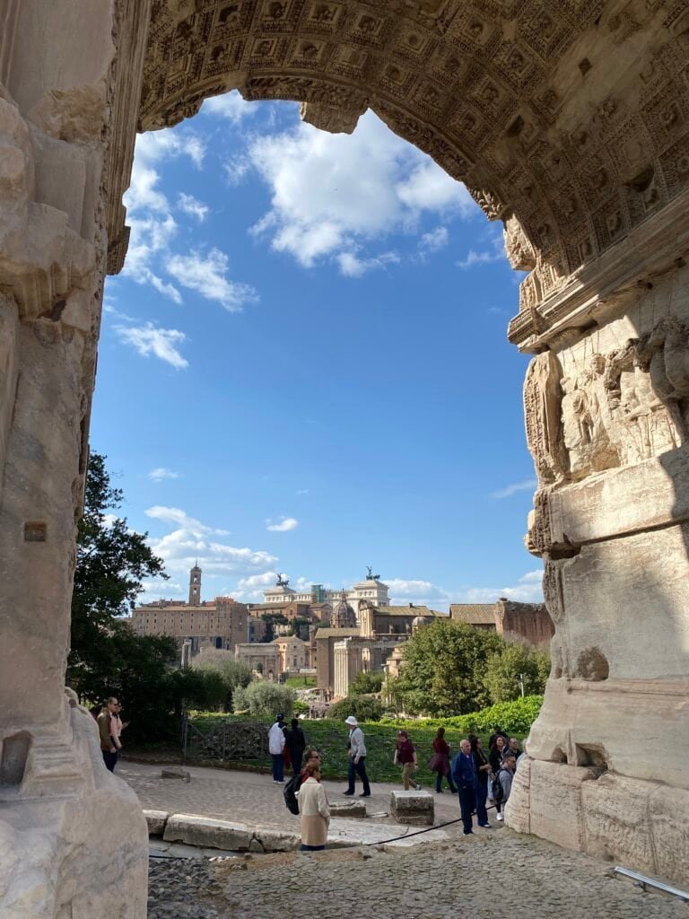 Ancient Roman arch framing a view of historic Pompeii ruins and Mount Vesuvius in the background, under a bright blue sky with scattered clouds.
