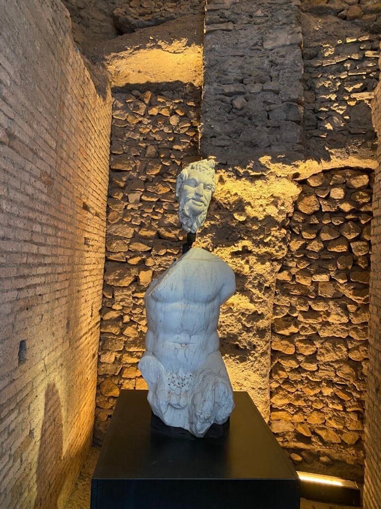 A classical Roman sculpture of a male figure with detailed musculature, displayed against a textured stone wall illuminated by warm lighting.