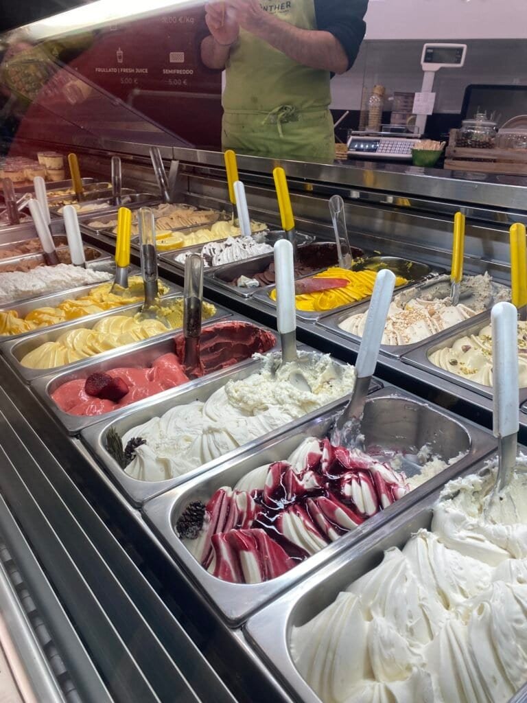 Colorful array of gelato flavors in metal containers, with various toppings and swirls, ready for serving at an ice cream parlor.