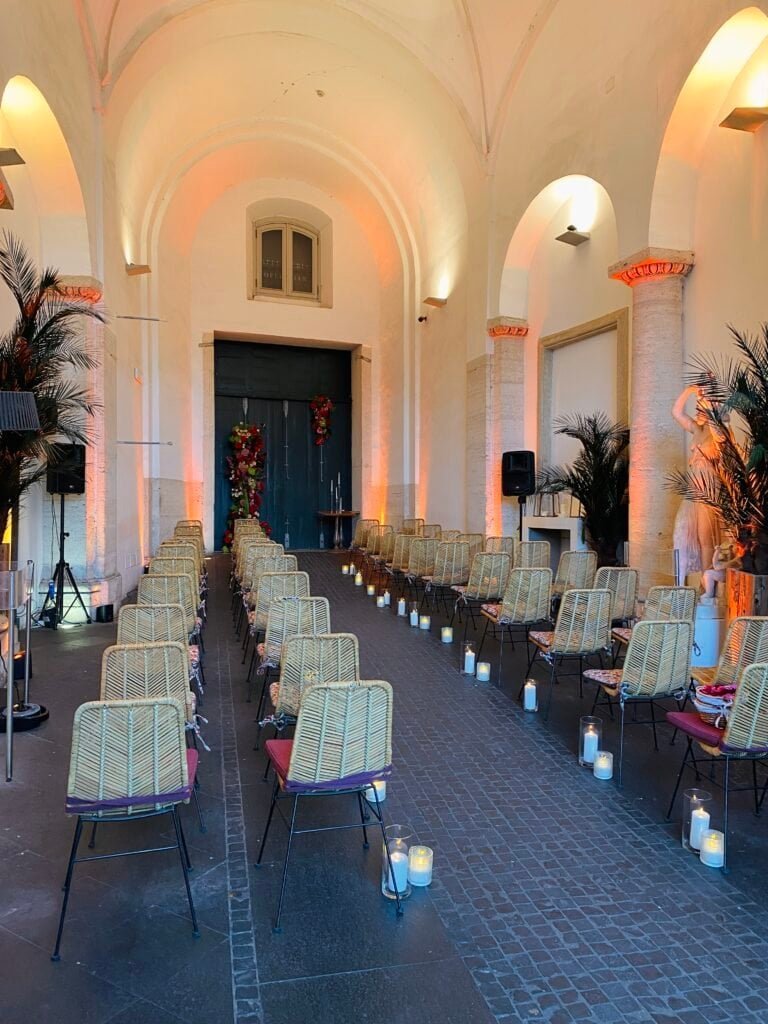 Elegant indoor wedding ceremony arrangement with chairs, candles, and tropical plants inside a historic church with high vaulted ceilings.