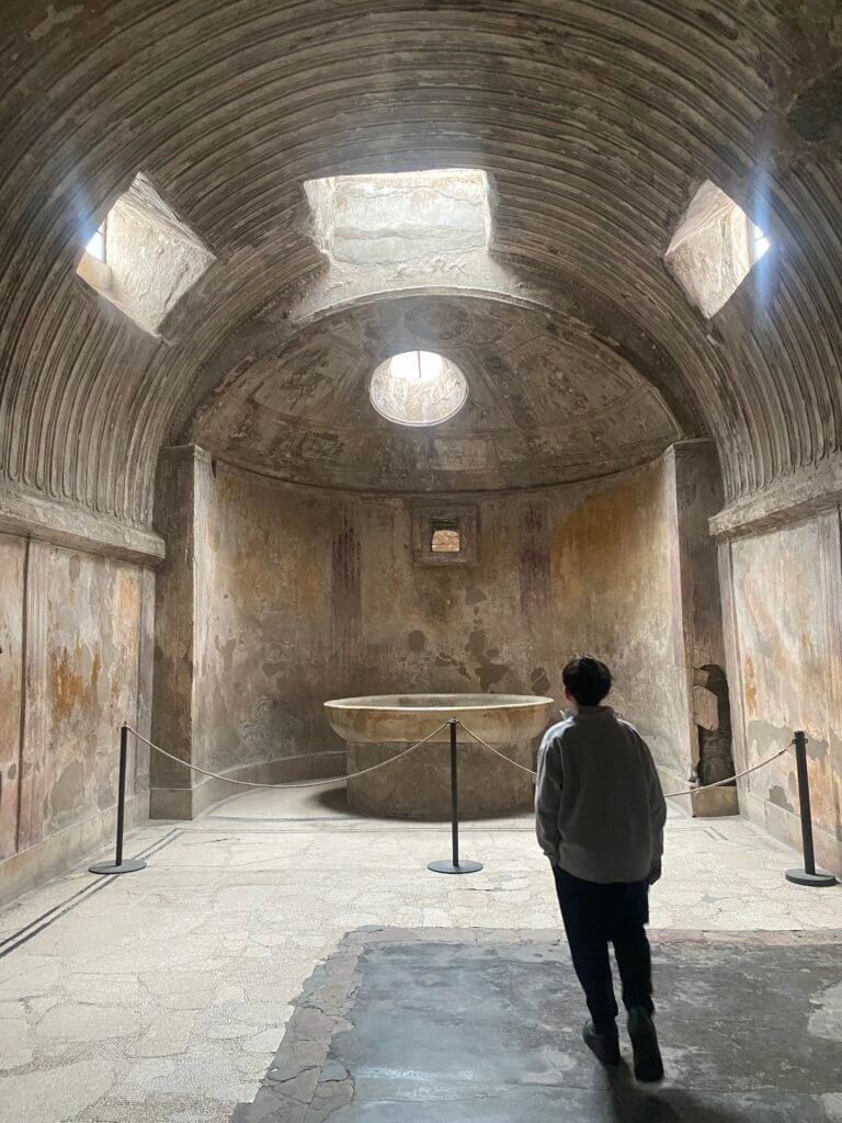 Interior view of an ancient Roman bathhouse with a central round pool, stone walls, and small windows for natural light, showcasing historical architecture.