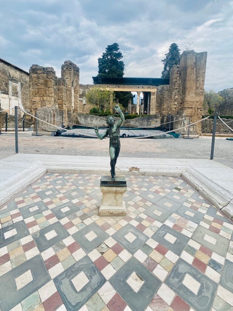 Ancient bronze Venus statue at Pompeii archaeological site, showcasing classical art amidst historic ruins, with a backdrop of ancient stone structures and lush greenery.