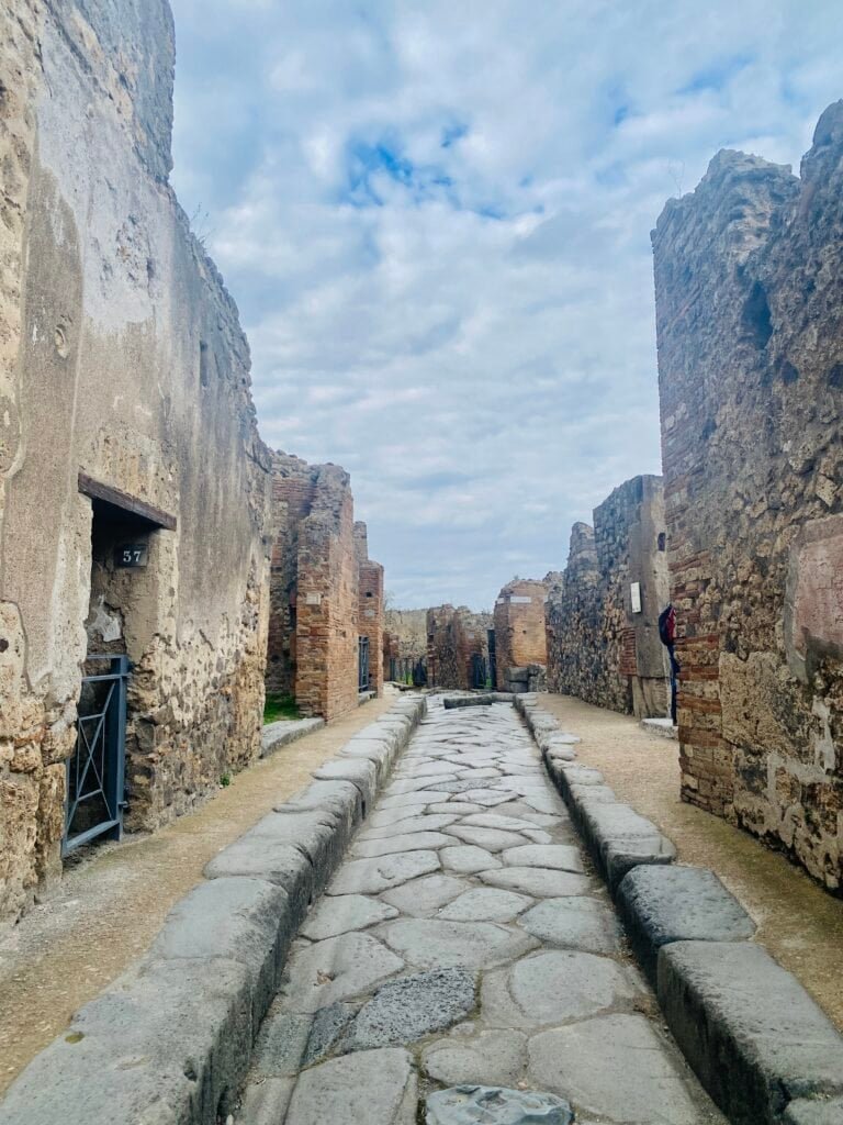 Ancient Roman street ruins in Pompeii with cobblestone pathway and stone walls, showcasing the historic architecture and archaeological significance of the site.