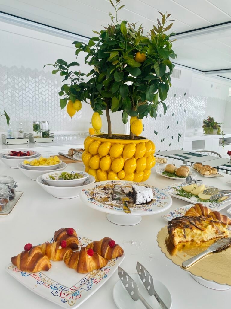 A vibrant lemon tree in a decorative yellow pot serves as a centerpiece on a white kitchen table, surrounded by various desserts and dishes, creating a lively and fresh atmosphere at Eden Roc positano italy.