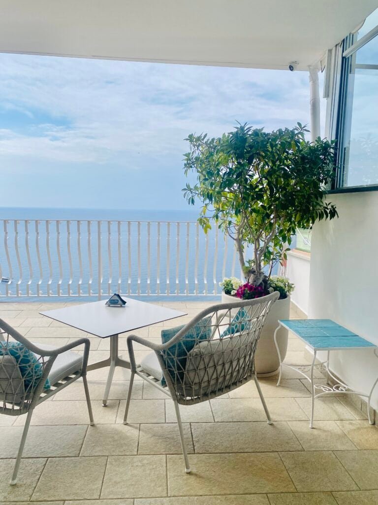 Bright balcony overlooking the ocean with seating and lush potted plant, capturing a serene seaside atmosphere.