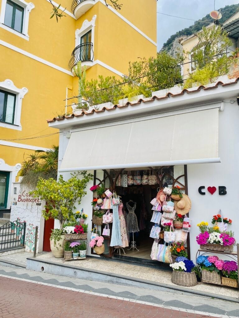 A quaint shop in Positano Italy showcasing vibrant clothing, accessories, and floral arrangements outside, set against a bright yellow building with lush greenery on the upper terrace.