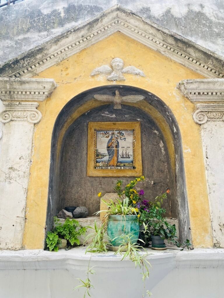 A colorful shrine in Italy featuring a religious icon, surrounded by potted plants and flowers, set against a yellow and gray wall with classical architectural details.