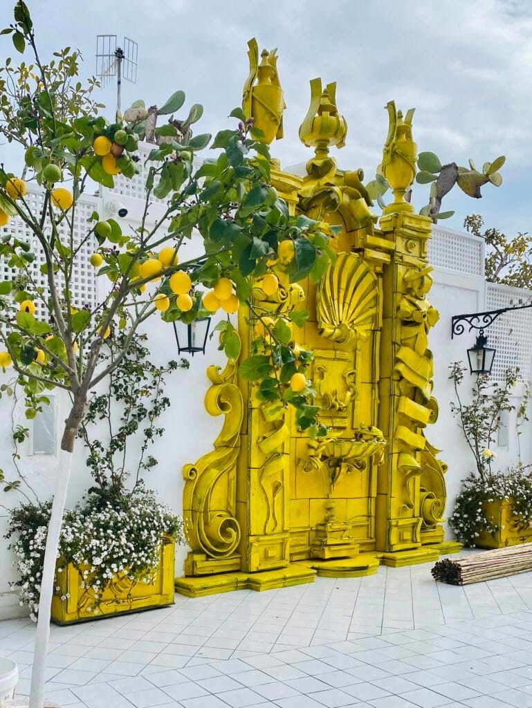 A bright yellow ornate wall installation with intricate details, set against a white background with potted plants and a lemon tree nearby.