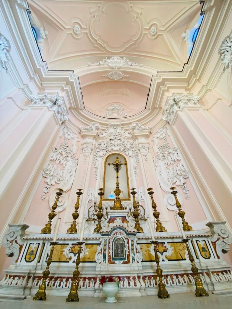Ornate Baroque church interior with intricate stucco details, pastel pink walls, and a gilded altar with candles, showcasing Italian religious architecture in Sorrento Italy.