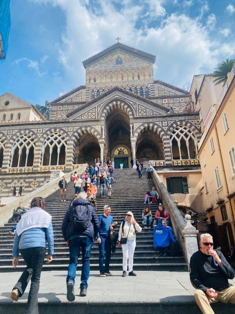 Tourists ascending the historic steps of a medieval cathedral in Amalfi Italy, showcasing intricate architecture and vibrant city life under a bright blue sky.