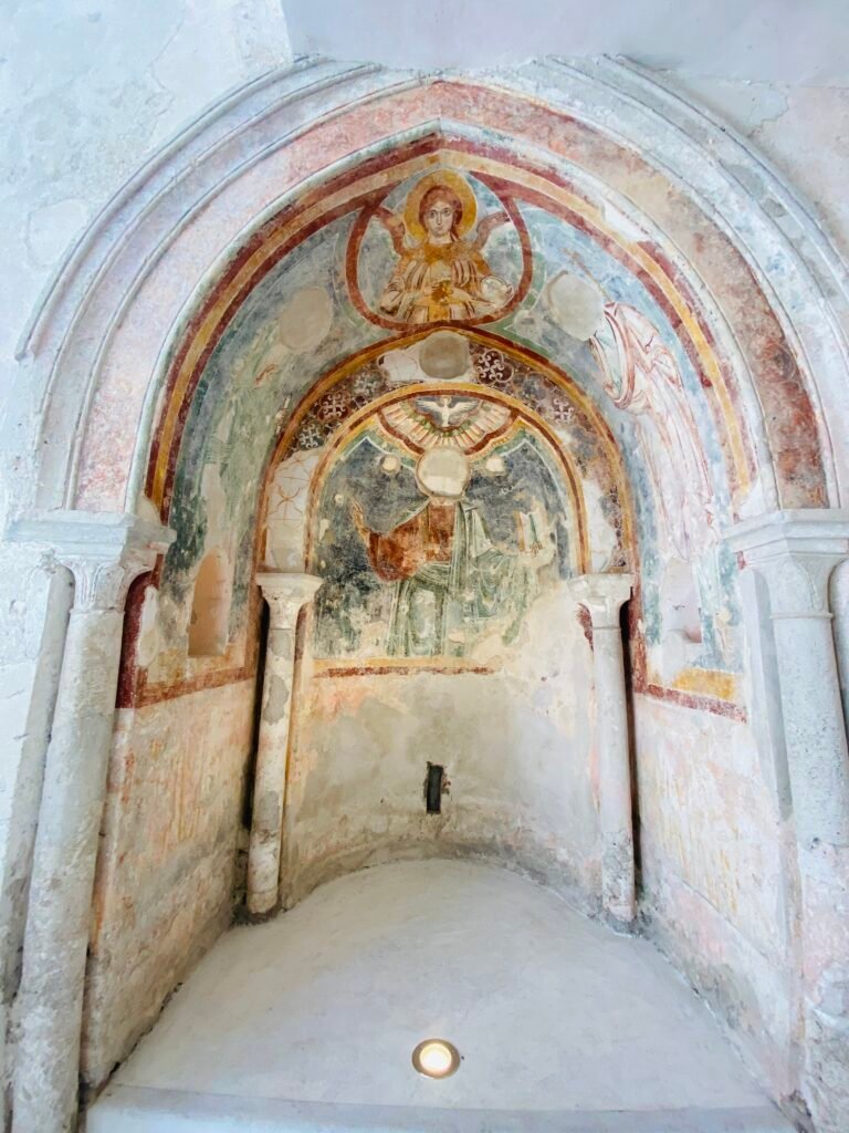 A historic fresco depicting a saint with a halo inside a small chapel in Italy. The artwork features vibrant colors and religious symbolism, showcasing Italy's rich cultural and spiritual heritage.