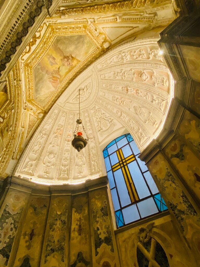 A detailed view of a church's ornate ceiling and stained glass window, highlighting intricate artwork, gold accents, and religious symbolism within a historic sacred space.