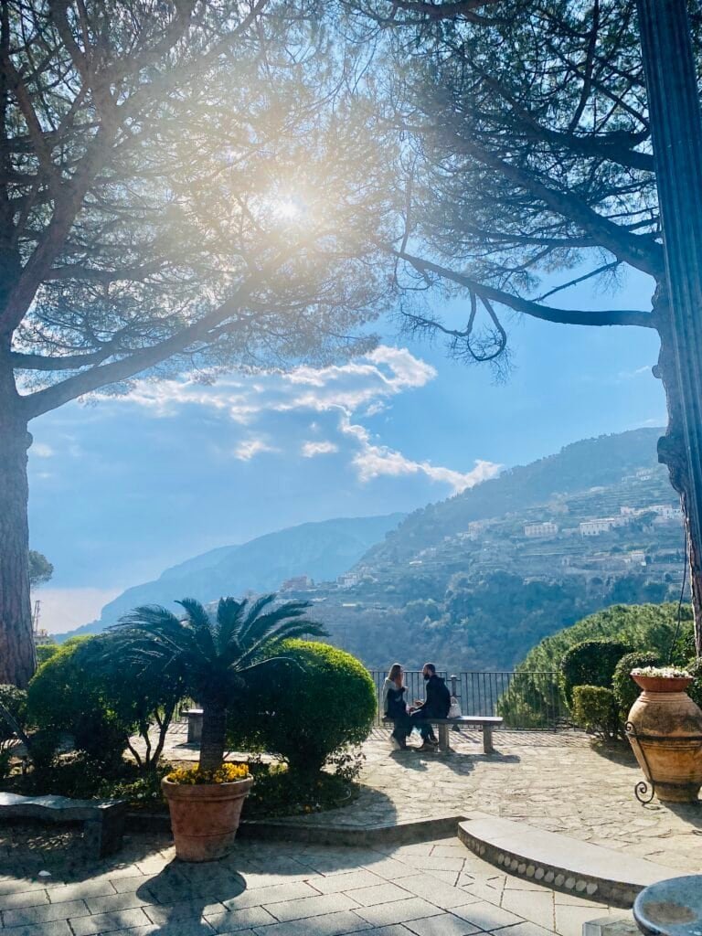 An idyllic scene featuring a sunny day in Italy with lush greenery, a clear blue sky, and a peaceful coastal landscape. Two people sit on a bench enjoying the view of the mountains and sea.