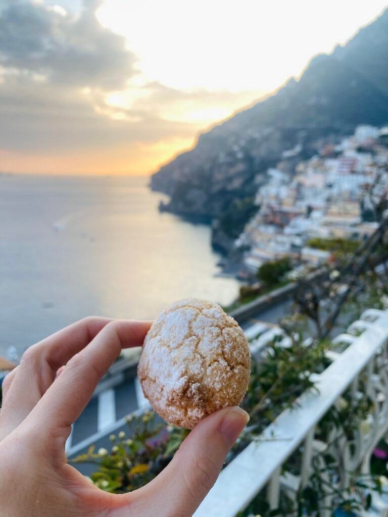 A hand holding a traditional Italian pastry with a scenic Amalfi Coast view at sunset in the background.