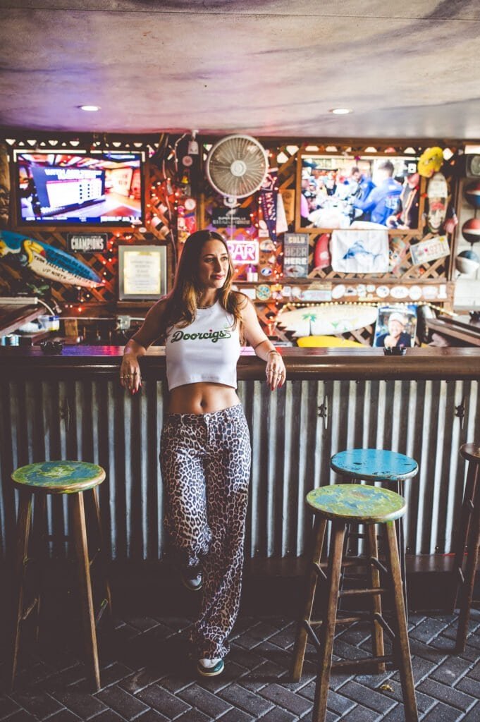 A woman with long hair, wearing a white crop top and leopard print pants, standing at a bar counter in a lively, colorful bar with neon signs and TV screens in the background.