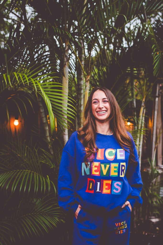 Smiling woman in a blue sweatshirt with "ICE NEVER DIES" text, standing among lush tropical plants in an outdoor setting, showcasing a cheerful and lively atmosphere.