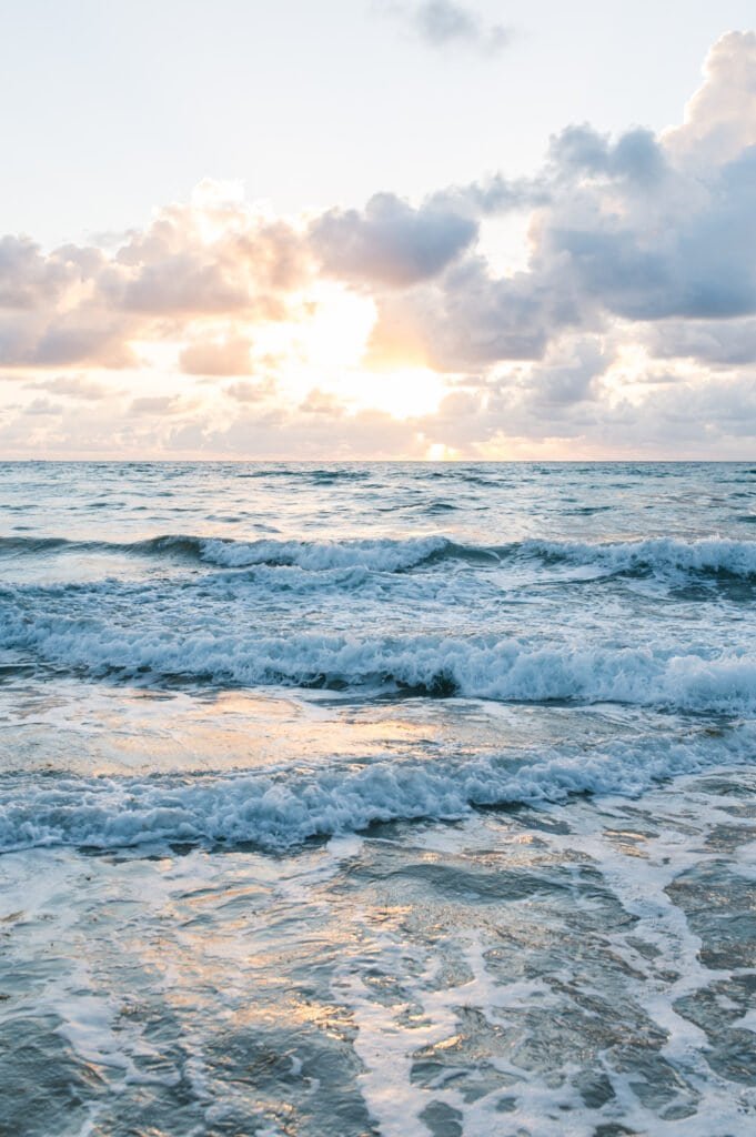 A serene sunset scene with the sun partially hidden behind clouds over the ocean, casting a warm glow on the waves and creating a peaceful, calming atmosphere.