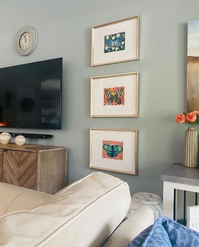 3 small butterfly paintings by Bonnie Sorsby in white mats and gold frames, stacked vertically on a narrow blue gray wall beside a TV