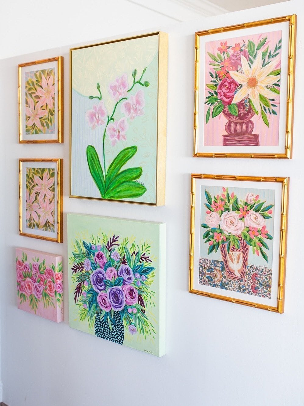 A gallery wall of floral originals by Bonnie Sorsby, including an orchid, roses and bouquets, mixed between gold bamboo frames and unframed canvases