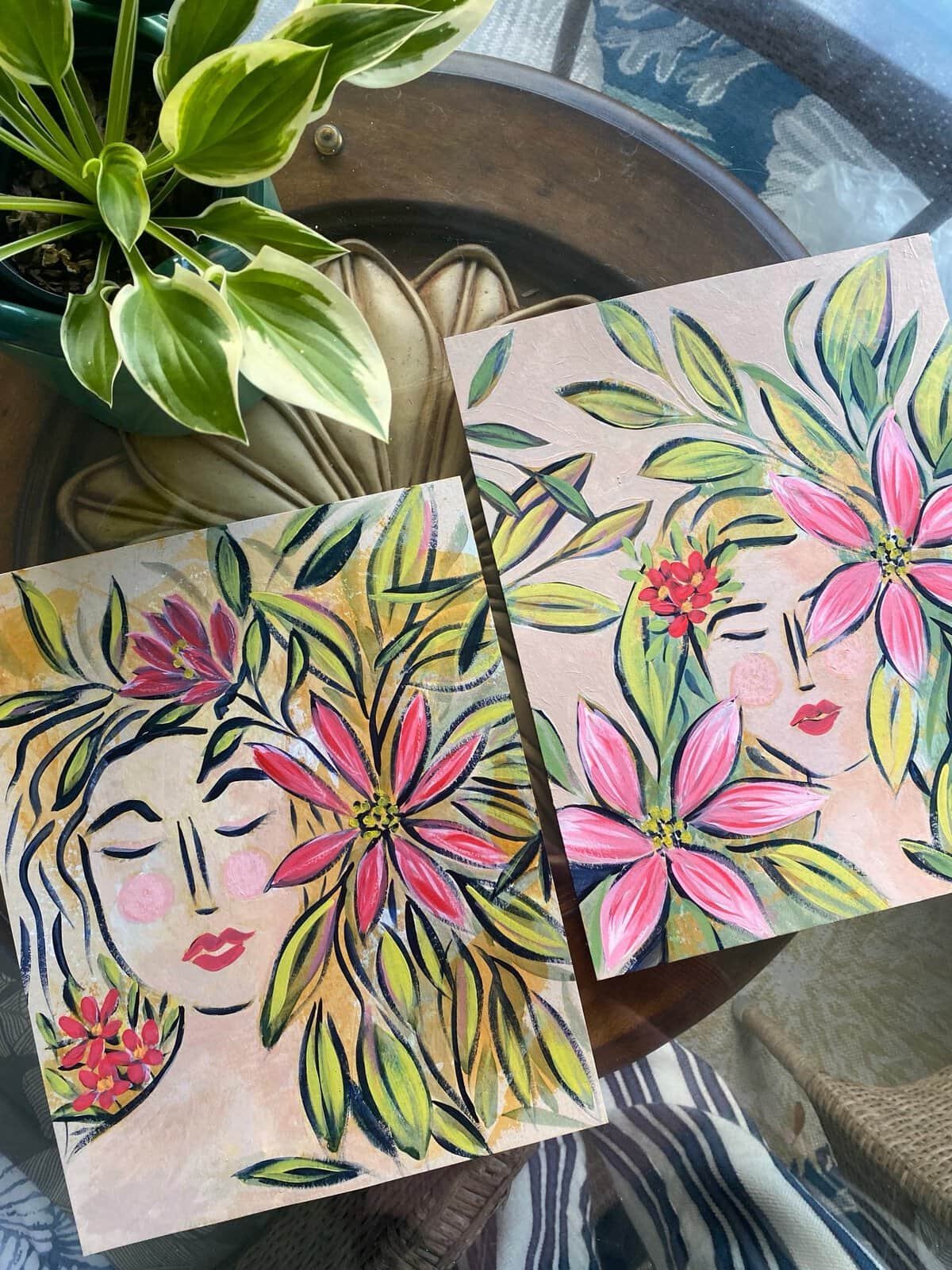 Tuned In and Tuned Out, a pair of original face paintings by Bonnie Sorsby with closed eyes, pink and red flowers and leafy hair, shown together on a wood table beside a hosta plant